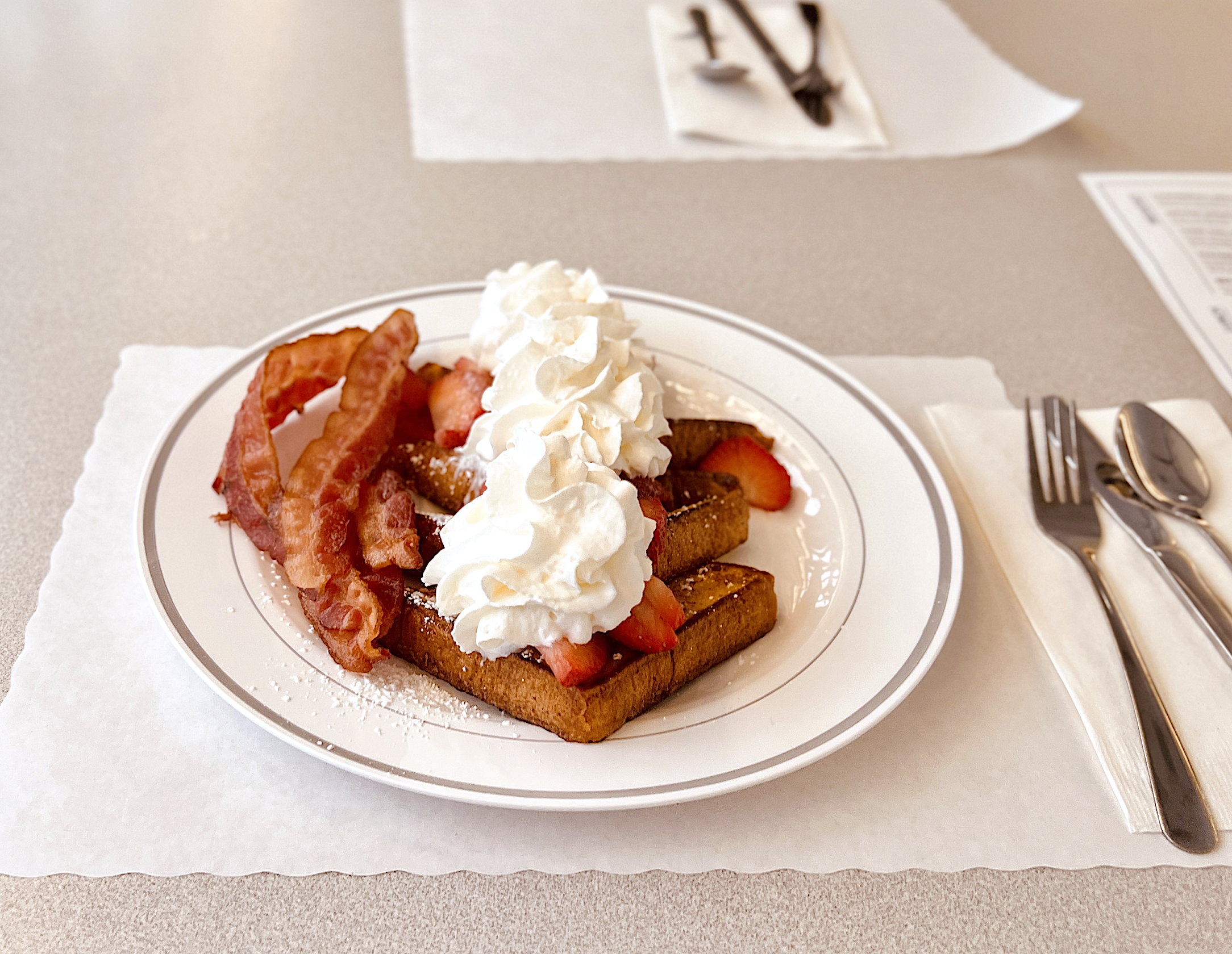 Ein Teller mit French Toast, garniert mit Erdbeeren und sehr, sehr viel Schlagrahm. Links daneben, vier Scheiben kross gebratenen Speck