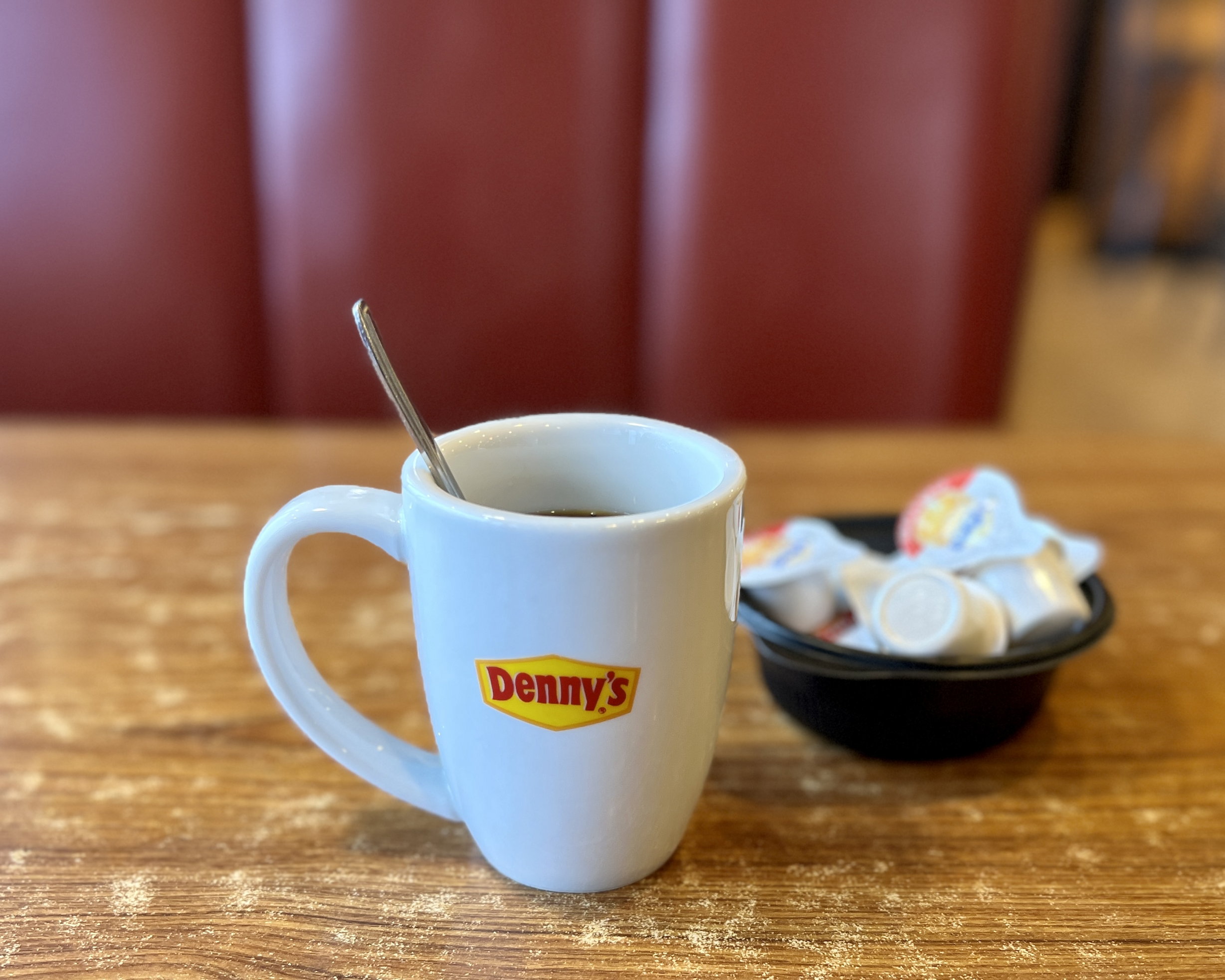 Eine Tasse Kaffee von Denny's in Grossaufnahme, daneben ein Schälchen mit Kaffeerahm. Dahinter, unscharf, das typische weinrote Kunstleder der Sitzbänke.