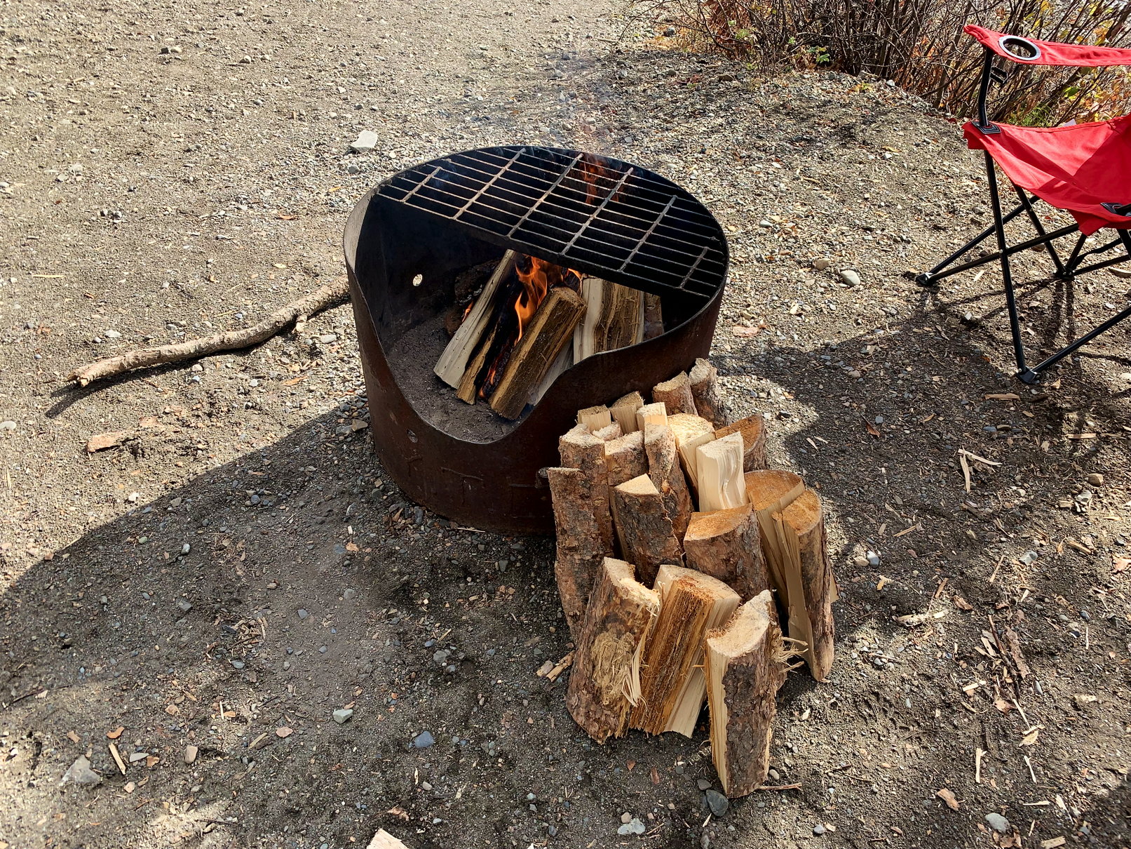 Das Feuer im Grill brennt, genügend Holz lagert daneben, im Hintergrund ein Campingstuhl