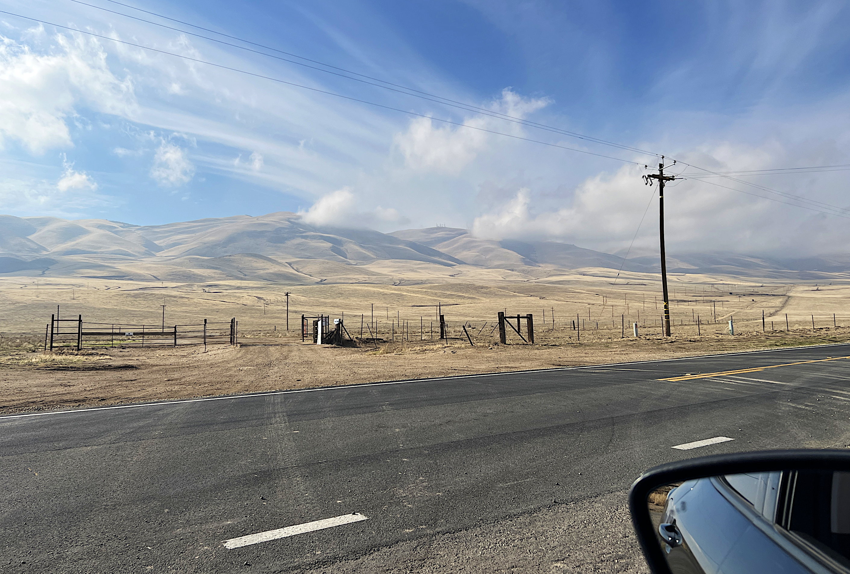 Aus dem Autofenster, man sieht einen Teil des Rückspiegels. Gegenüber braune, vertrocknete Wiesen und Hänge. Die Hügel sind teilweise in den Wollken. Der Himmel ist milchig blau.