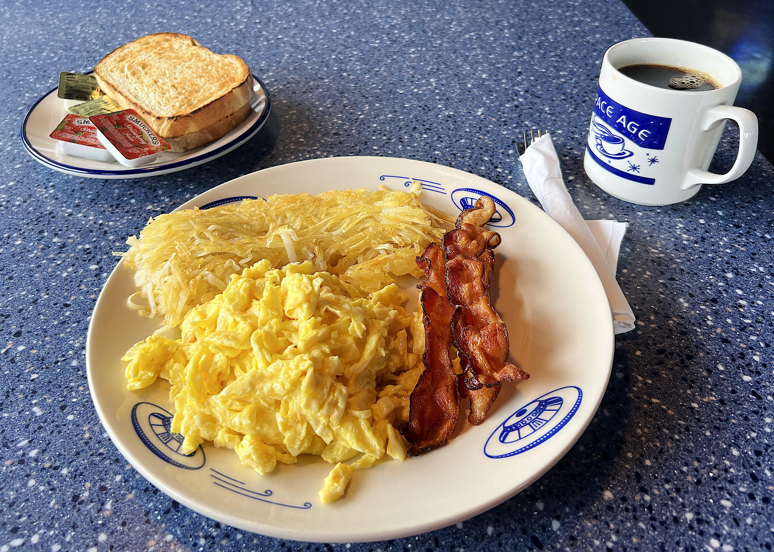 American Breakfast. Zwei (grosse!) Rühreier, zwei Stück Speck, kross gebraten. Dazu Rösti, Kaffee und Toast.