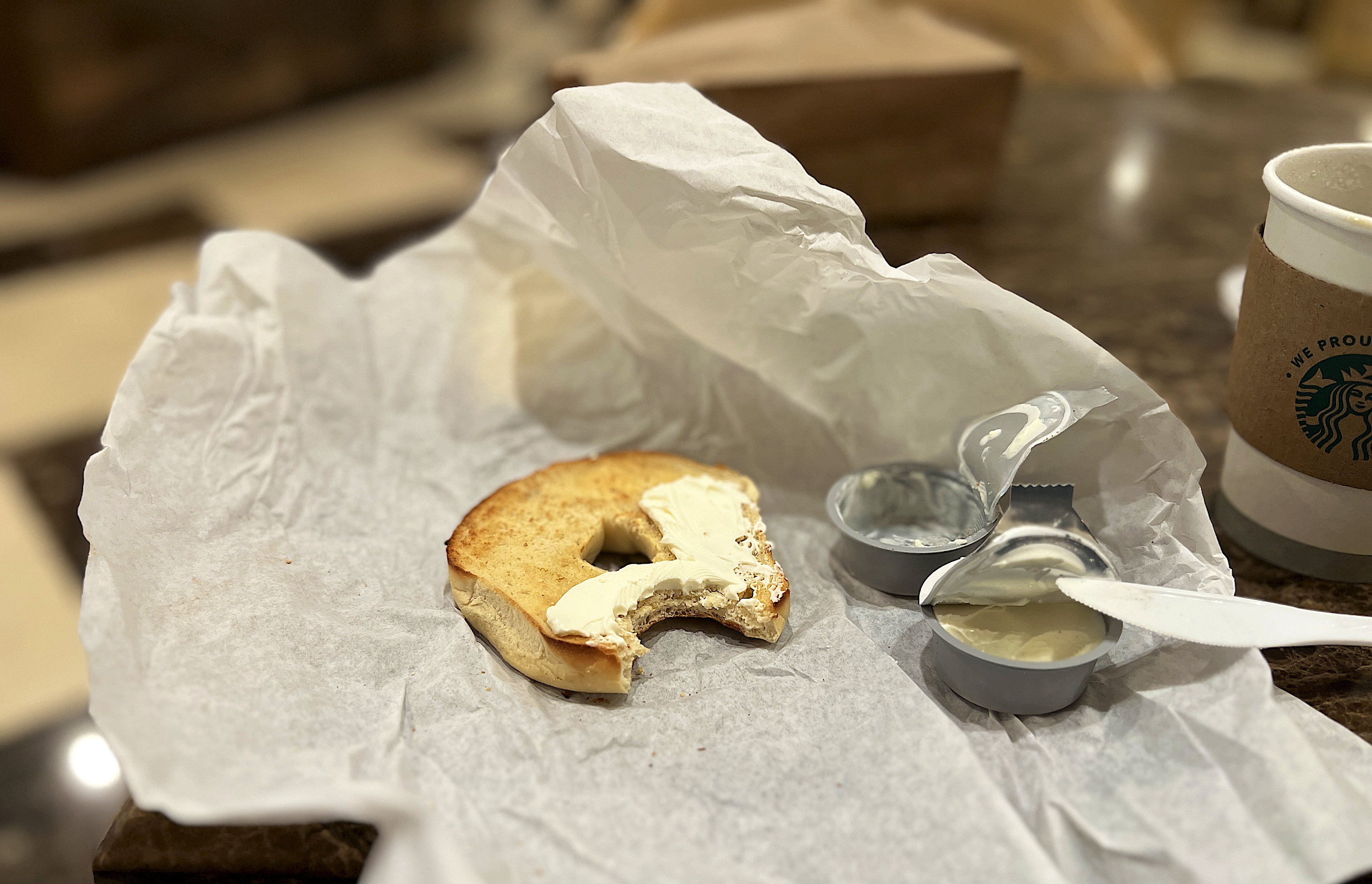 Auf dem Einwickelpapier liegt ein angebissener, getoasteter Bagel mit Cream Cheese. Daneben die leeren Cream Cheese Döschen.