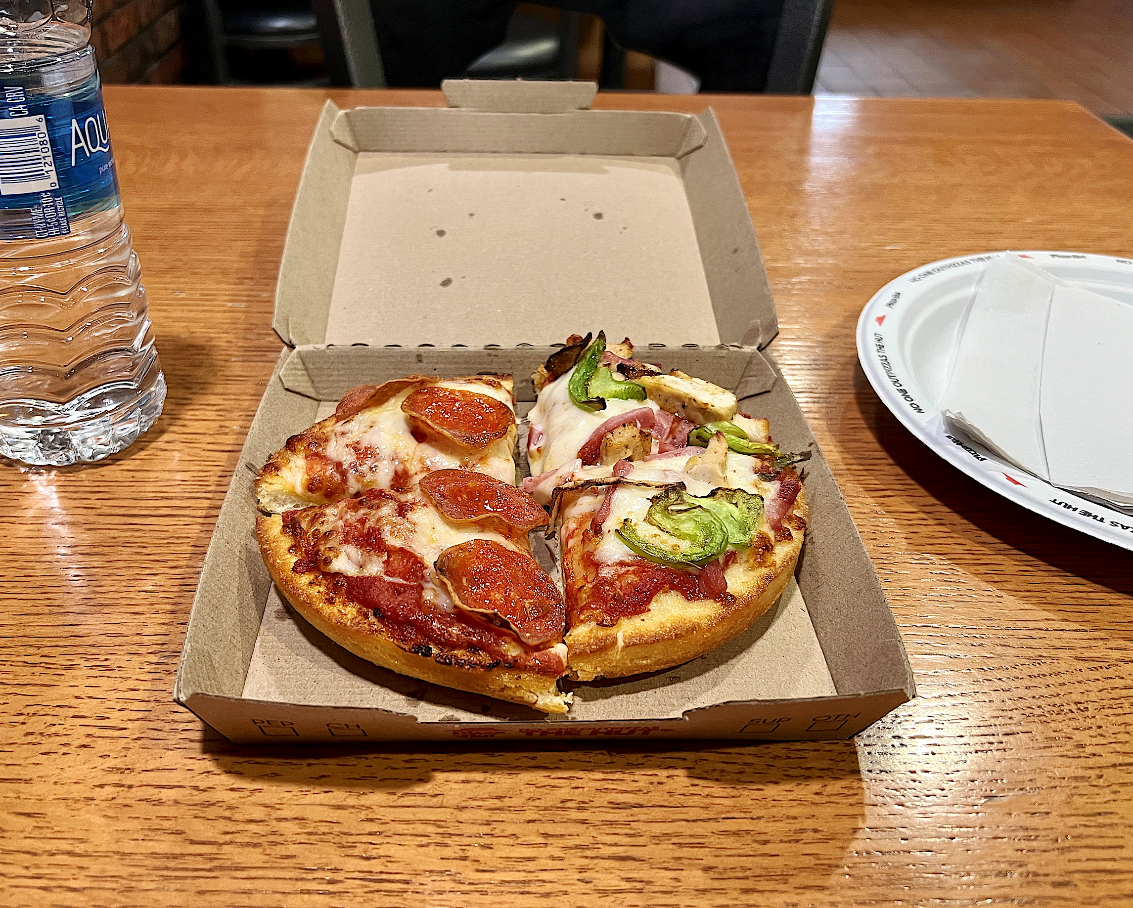 Eine kleine, rund 20cm durchmessende, dicke Pizza in der geöffneten Kartonschachtel. Auf einer Seite mit pikanter Salami, auf der anderen Seite mit Gemüse und Schinken. Hätte Hawai'i sein sollen. Daneben steht eine Flasche Wasser.