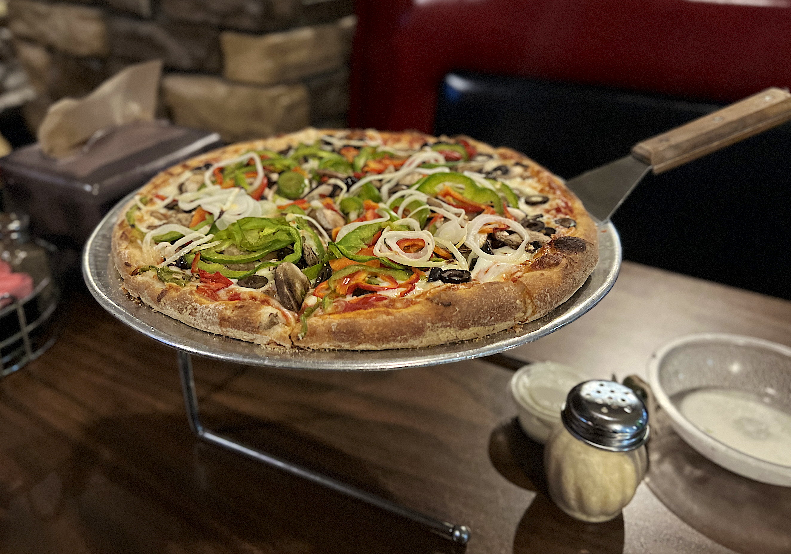 Eine etwas mehr als tellergrosse Pizza auf einem Blech, das auf einem Ständer auf dem Tisch steht. Die Pizza ist sehr, sehr reichhaltig mit Paprikas, Oliven, Pilzen und Zwiebeln belegt.
