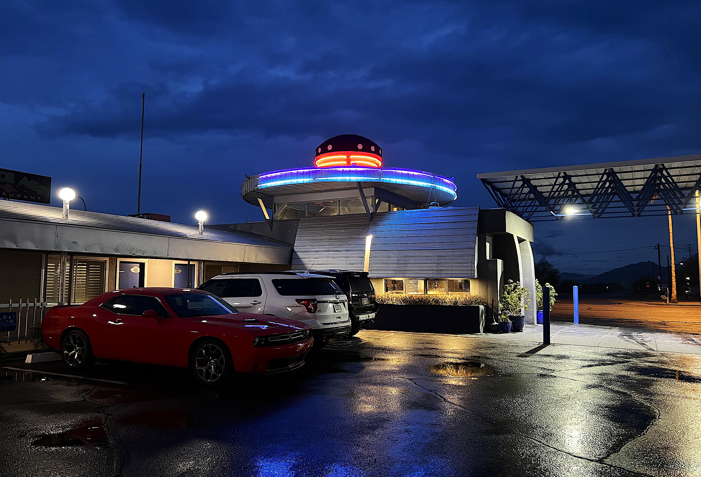 Blick auf das im nächtlichen Licht liegende Hotelgebäude mit dem markanten Aufsatz, der wie ein UFO aussieht. Das Licht spiegelt sich in den Pfützen auf dem Parkplatz, wo mehrere Autos parkiert sind.