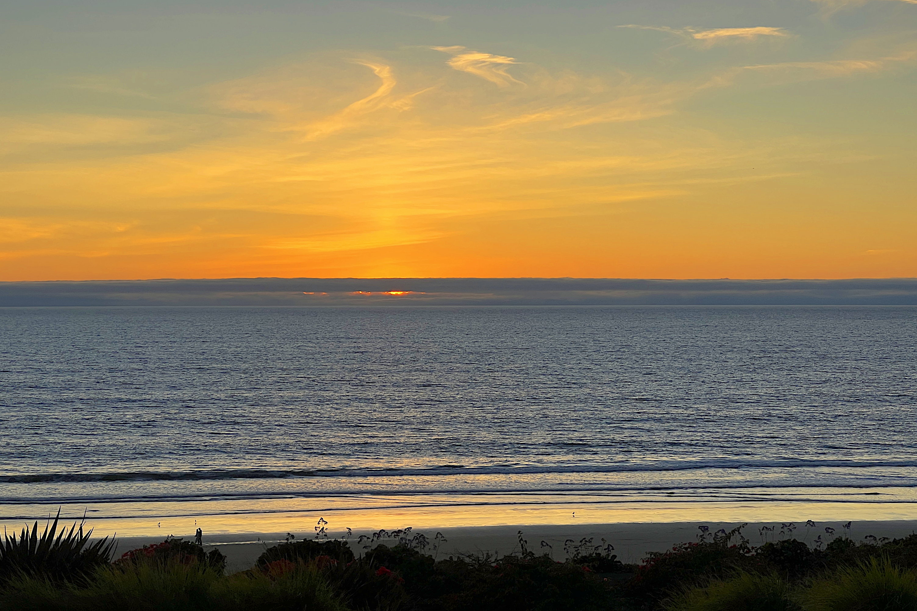 Wieder ein Sonnenuntergang über dem Pazifik. Die Sonne ist unter die Wolkenbank am Horizont gerutscht. Sie scheint aber ein klein wenig unter der Wolkenbank hervor. Der Himmel ist orange und die wenigen hohen Wolken werden von unten beleuchtet.