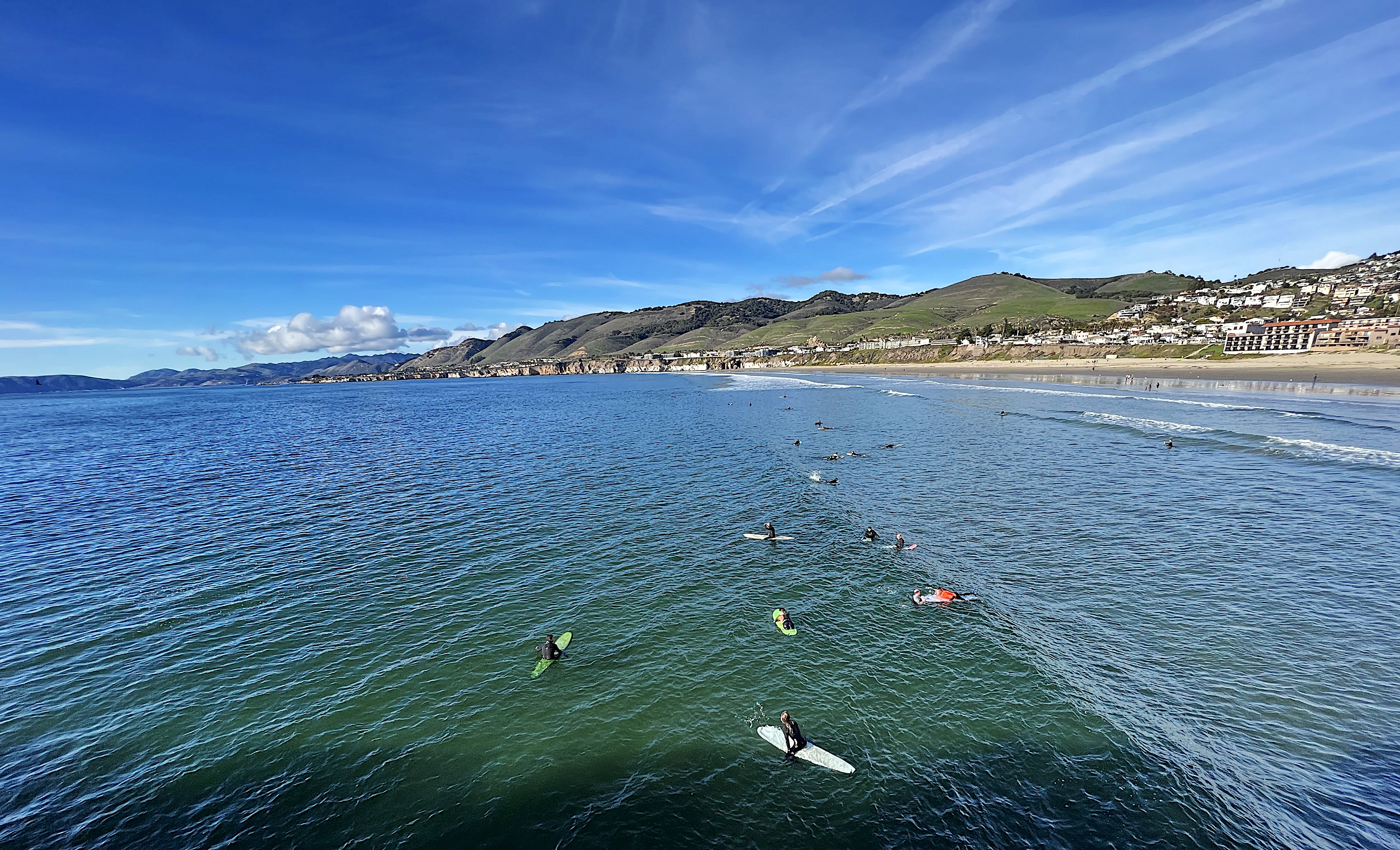 Ein Dutzend Surfer wartet in der Dünung auf den Brettern liegend oder sitzend auf die richtige Welle. Dahinter der Strand und die Klippen der Bucht.