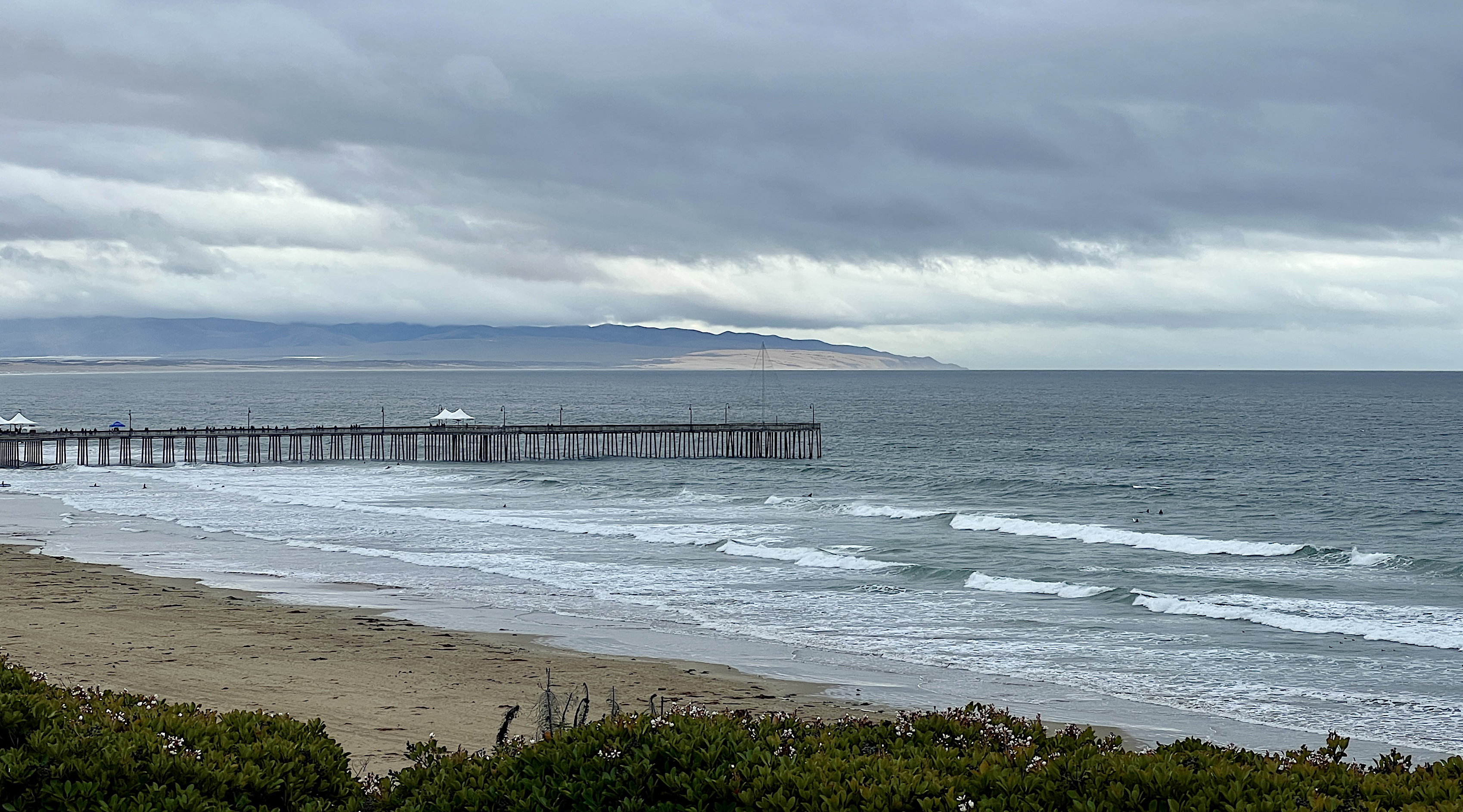 Der Strand mit Pier in Pismo Beach gegen 10 Uhr. Der Himmel ist grau, das Meer ziemlich aufgewühlt. Ein paar Unentwegte sind auf ihren Surfbrettern im Meer.