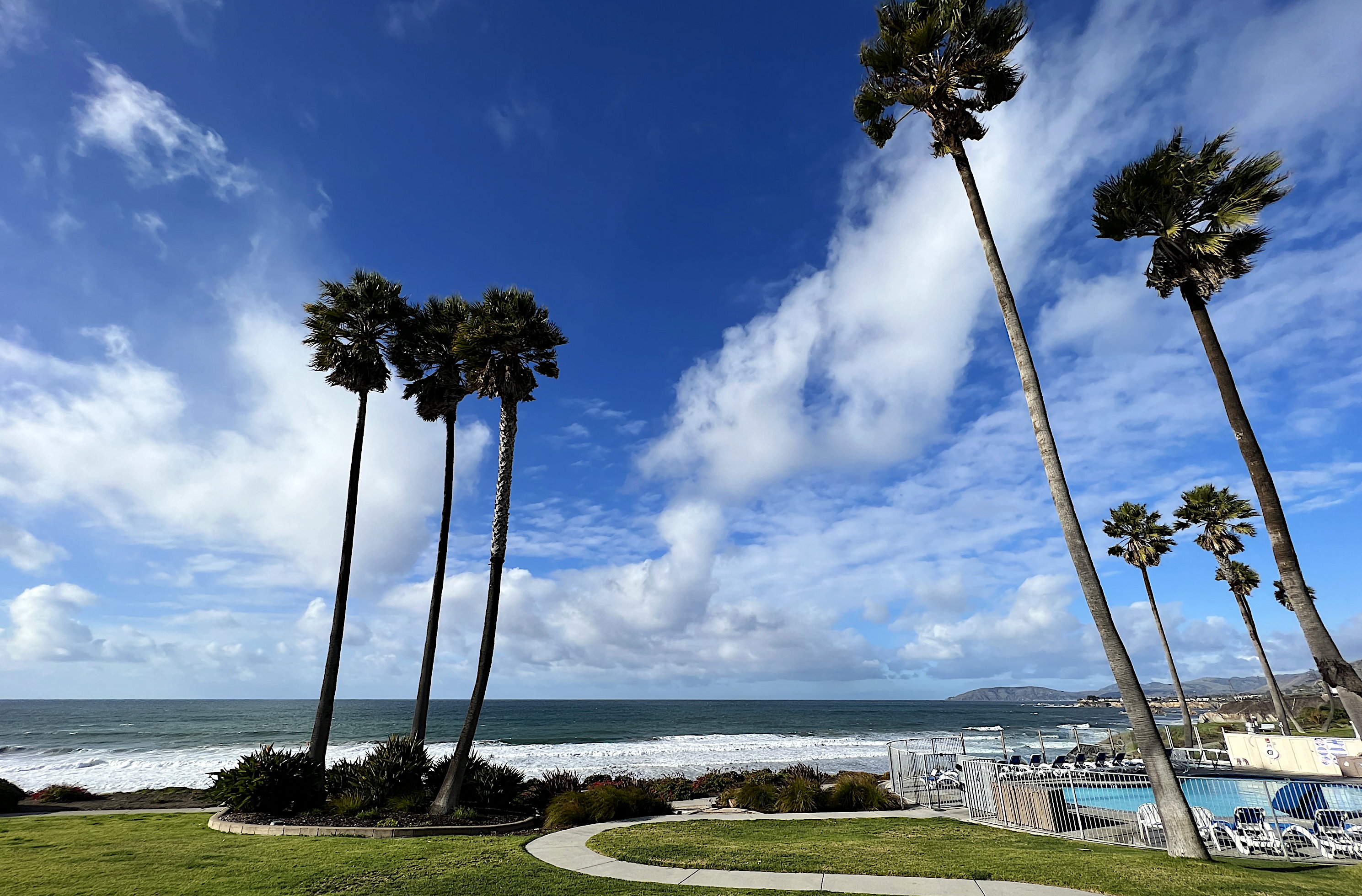 Blick vom Balkon des Hotels auf den Pazifik und Richtung Nordwesten. Blauer Himmel mit weissen Wolken, immer noch windig. Im Vordergrund die Palmen und der Pool.