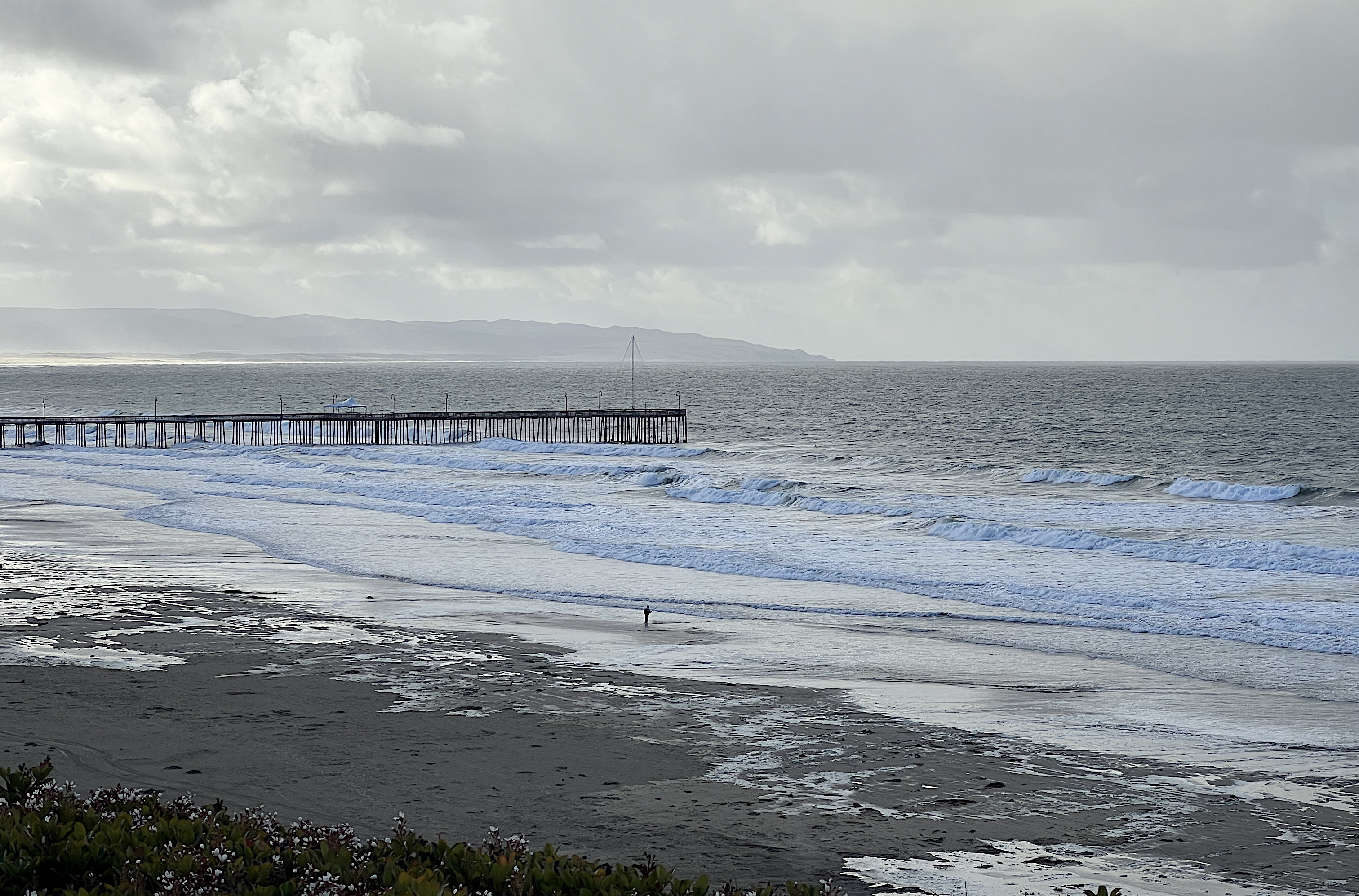 Blick vom Hotel zum Pier. Das Meer ist aufgewühlt und die Wellen werfen weisse Gischt. Wenige Leute sind am Strand zu sehen. Der Himmel ist grau.