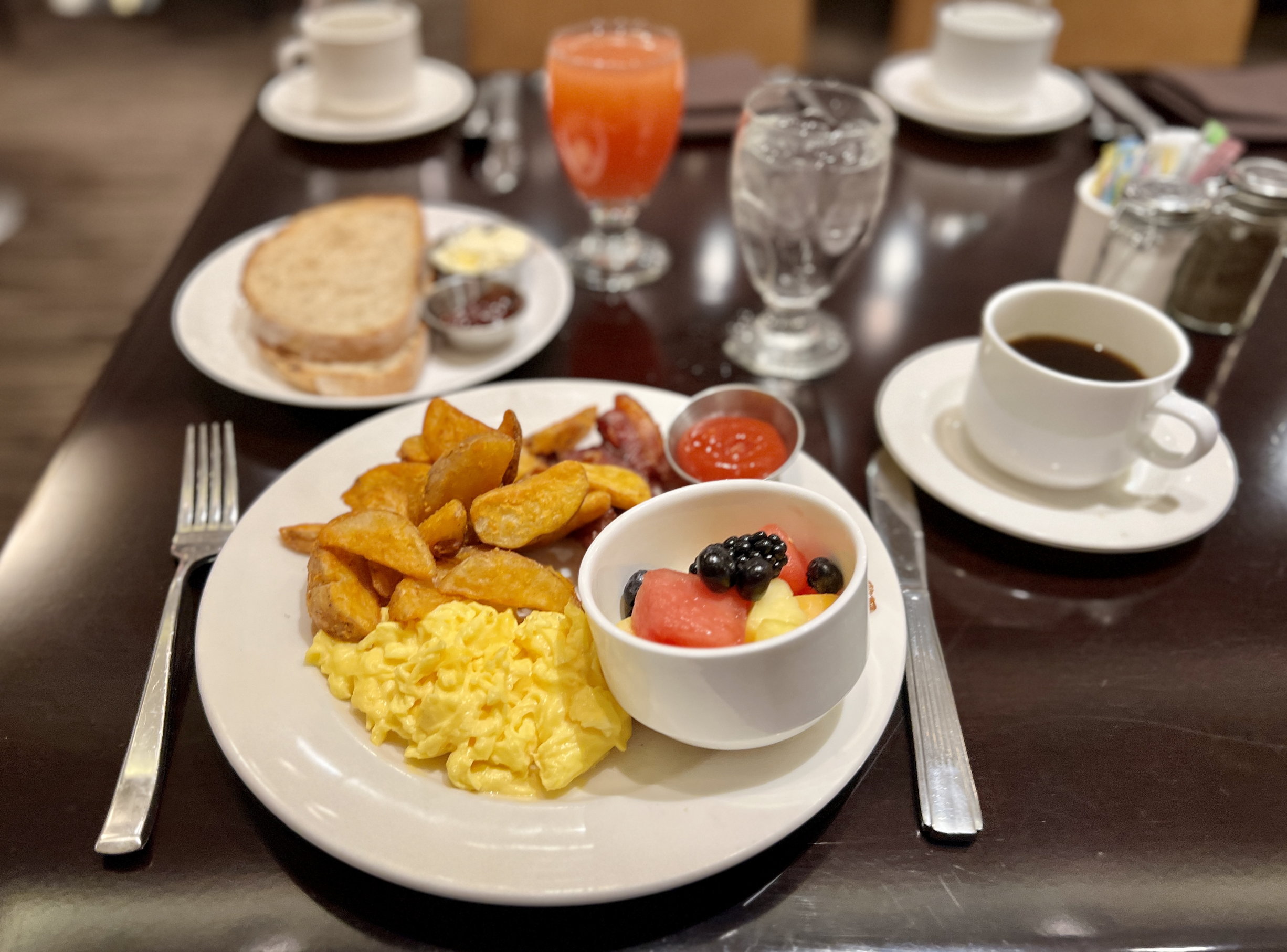 Ein reichhaltiges US-Frühstück mit Rührei und Kartoffeln, dazu Früchte und dahinter Kaffee und Toastbrot mit Konfitüre